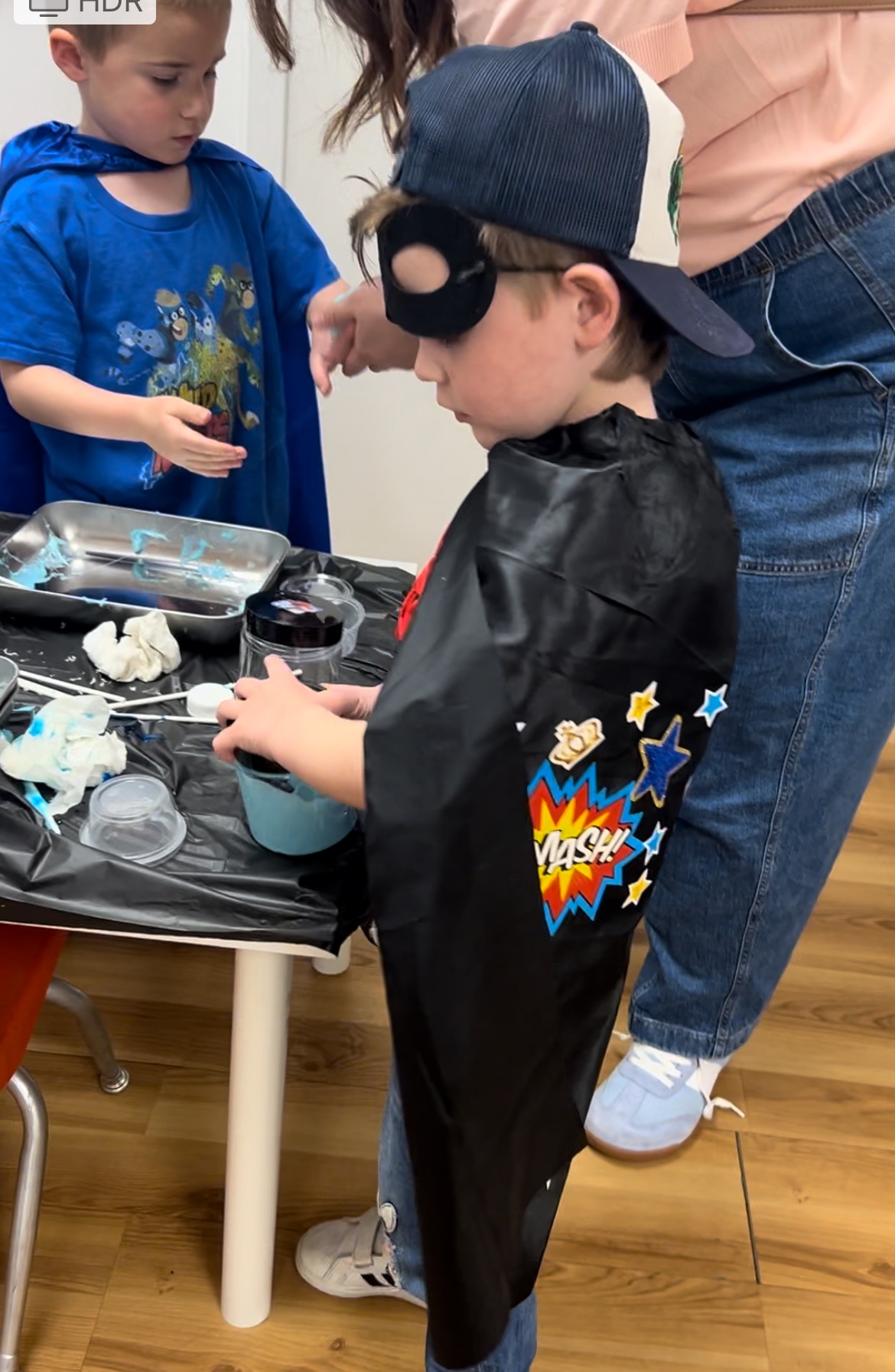 A boy in a superhero cape and mask doing a messy art project at a party